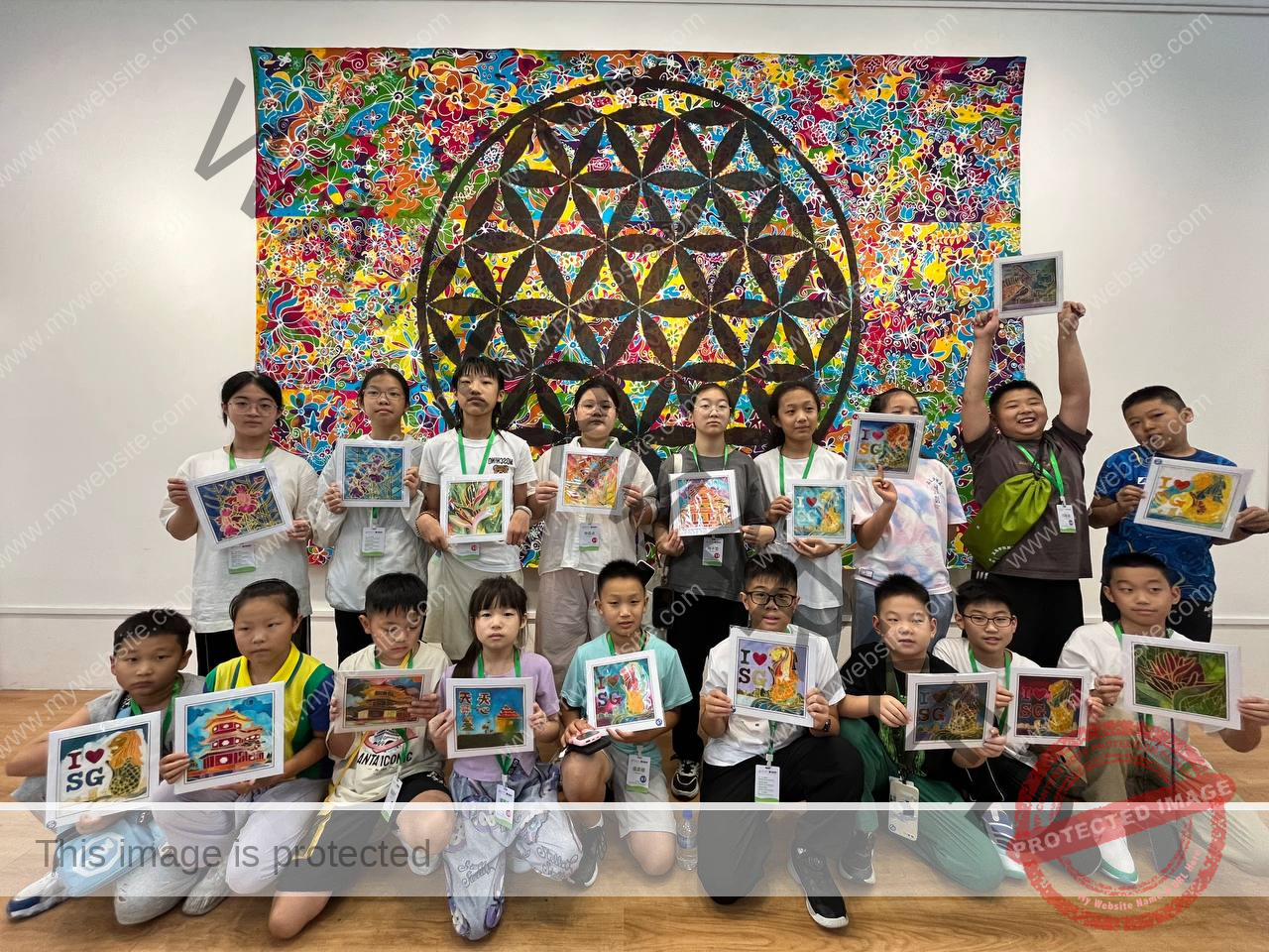 A group of children stand in front of a colorful geometric mural, proudly holding up their own artwork created during a Singapore Summer Immersion Program.