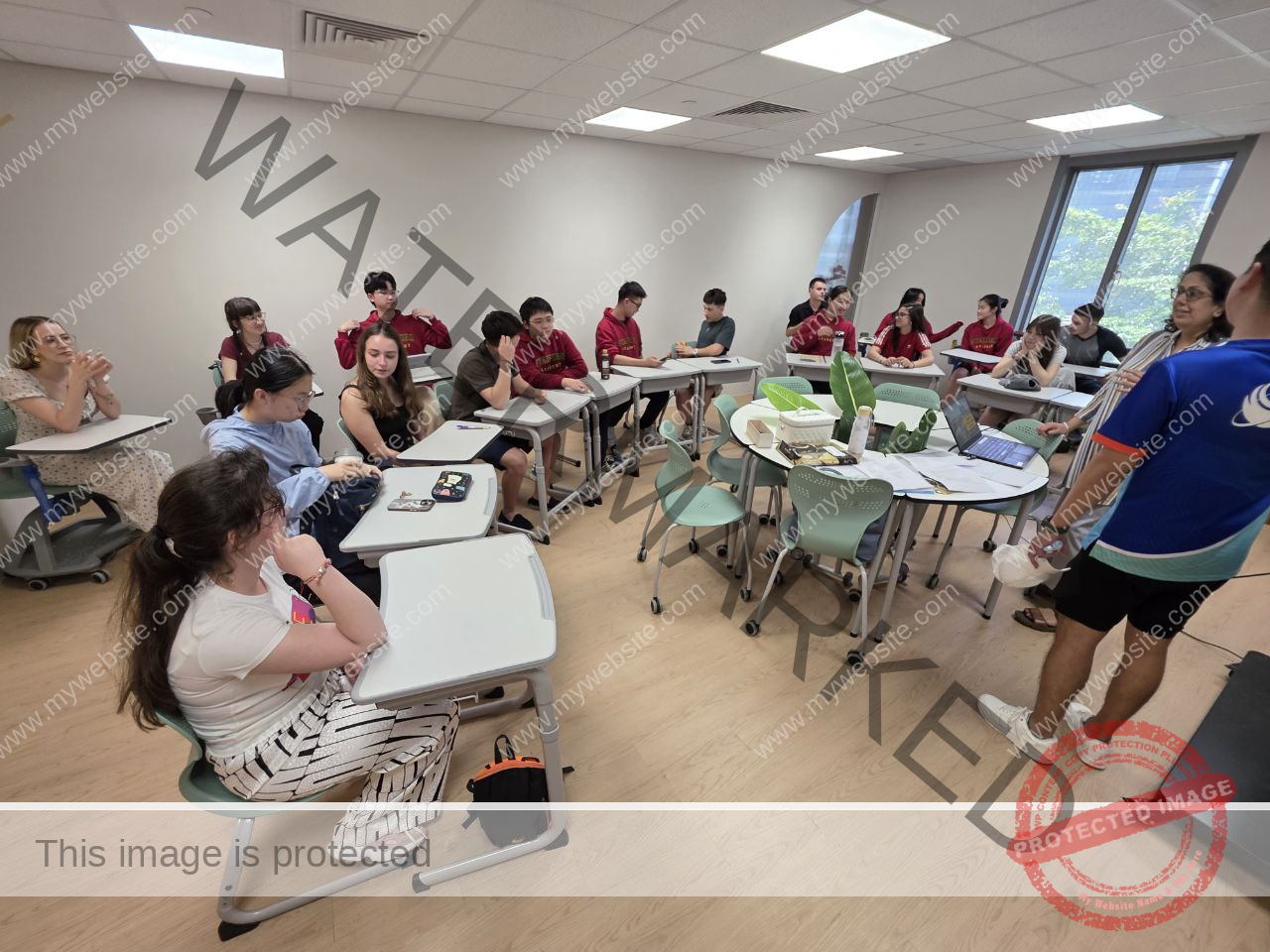 A teacher stands at the front of a classroom, addressing our students seated at individual desks arranged in a semi-circle during English summer camp.